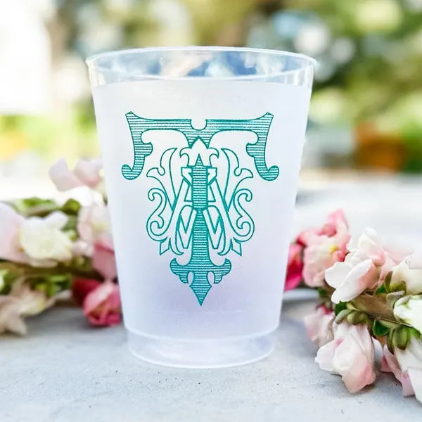 A white plastic cup with a green monogram design on it, placed on a table with pink flowers in the background.