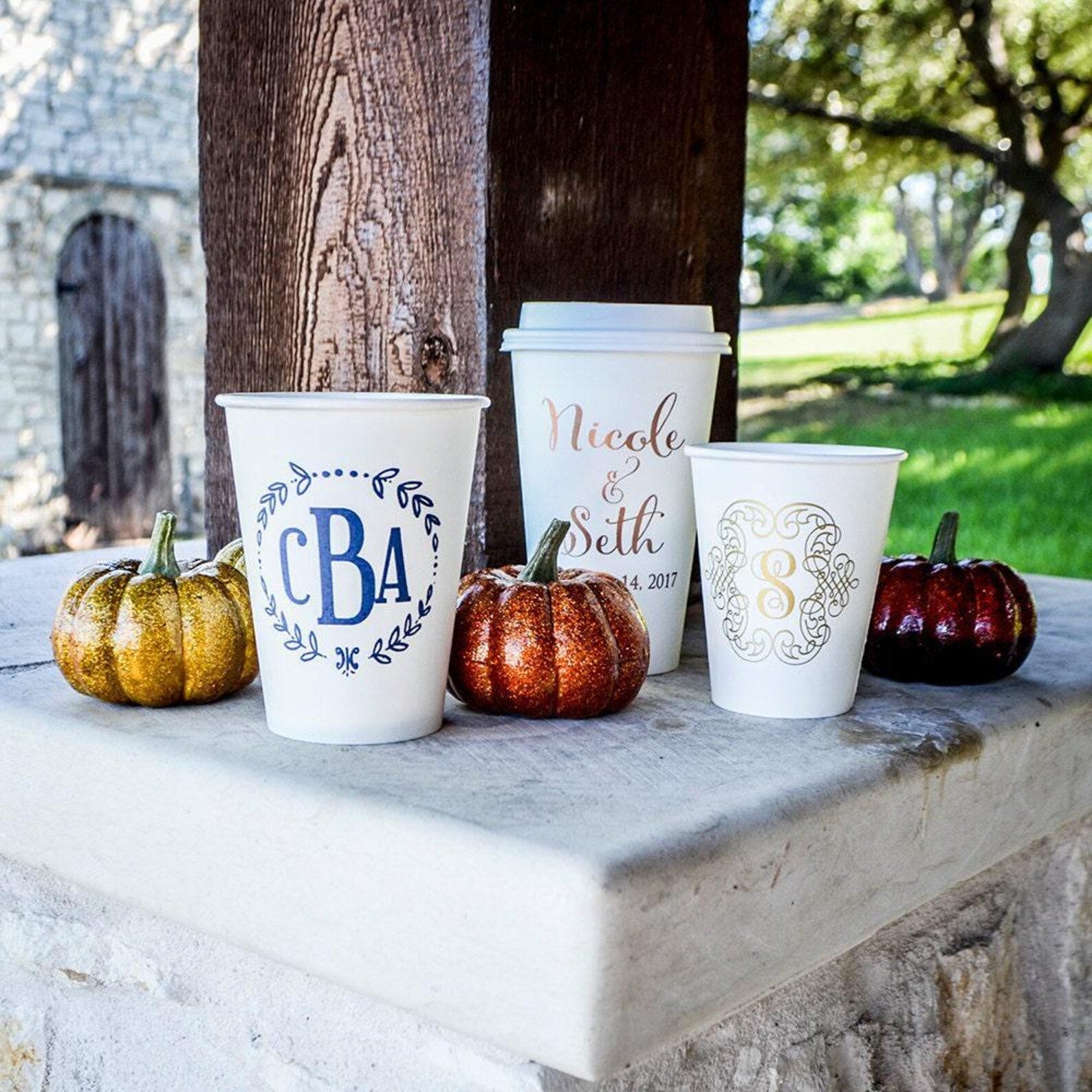 a white coffee cup with a personalized monogram, a white paper cup with a personalized monogram, and three small pumpkins with a gold and red glitter finish.