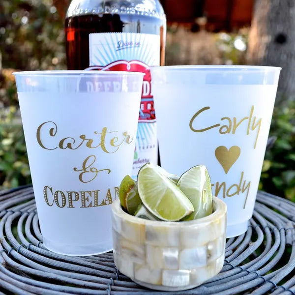 a close-up of a set of personalized cups, a bottle of alcohol, and a bowl of lime wedges, all placed on a woven surface.