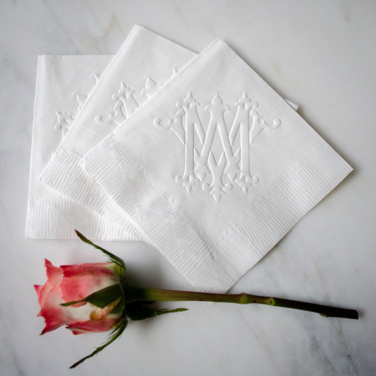 a pink rose with three white napkins with an ornate monogram design on them, placed on a marble surface.