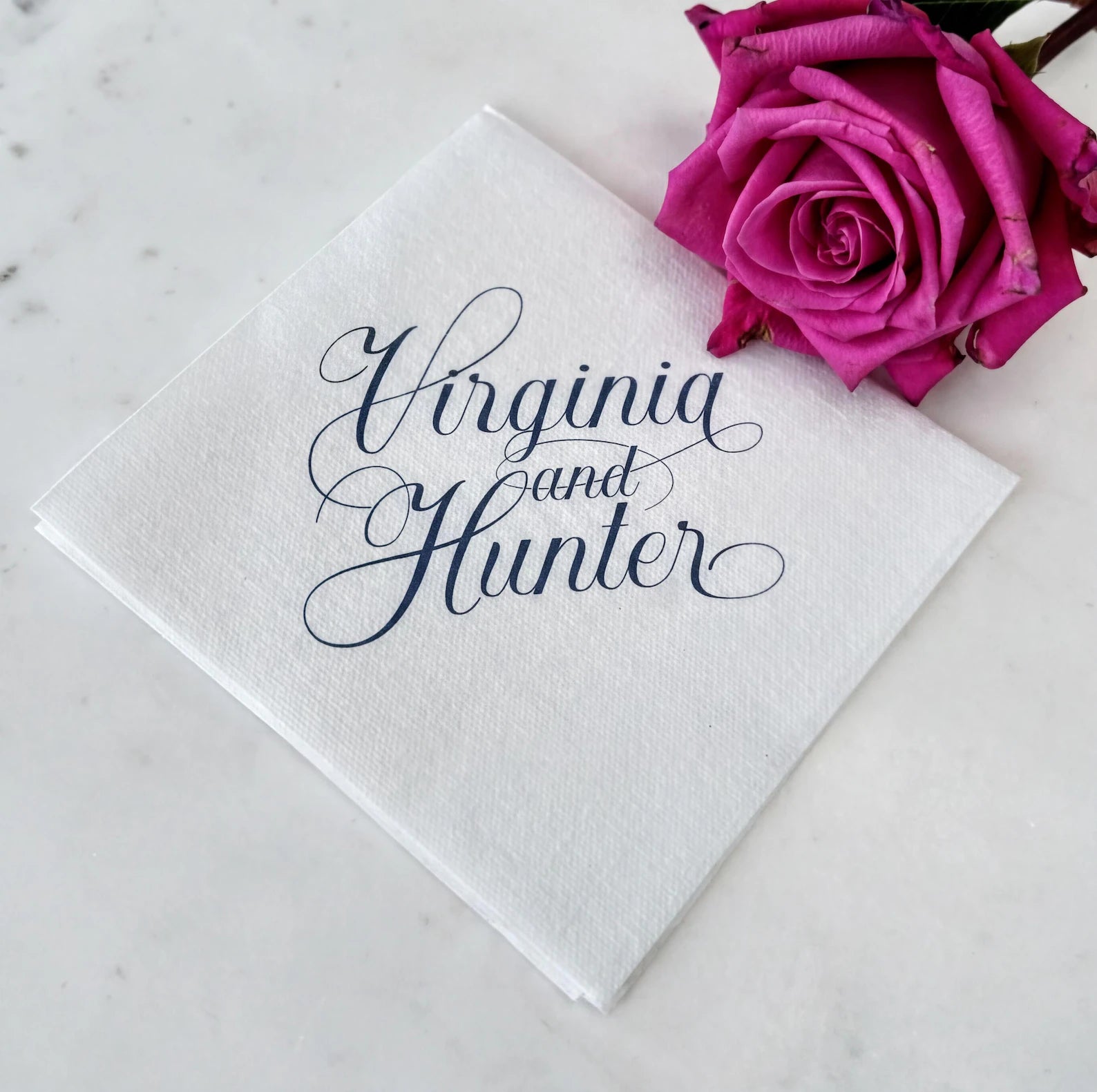 a white napkin with the text "Virginia and Hunter" written on it, and a single pink rose is placed next to the napkin.