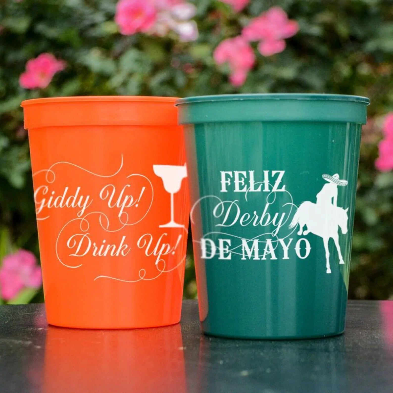 Two colorful plastic cups with text printed on them, placed on a table with pink flowers in the background.