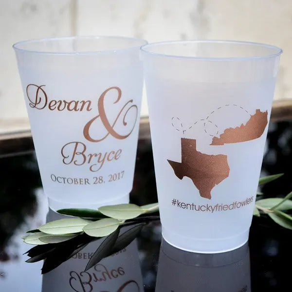 Two white plastic cups with printed text and graphics on them, placed on a reflective surface with some greenery.