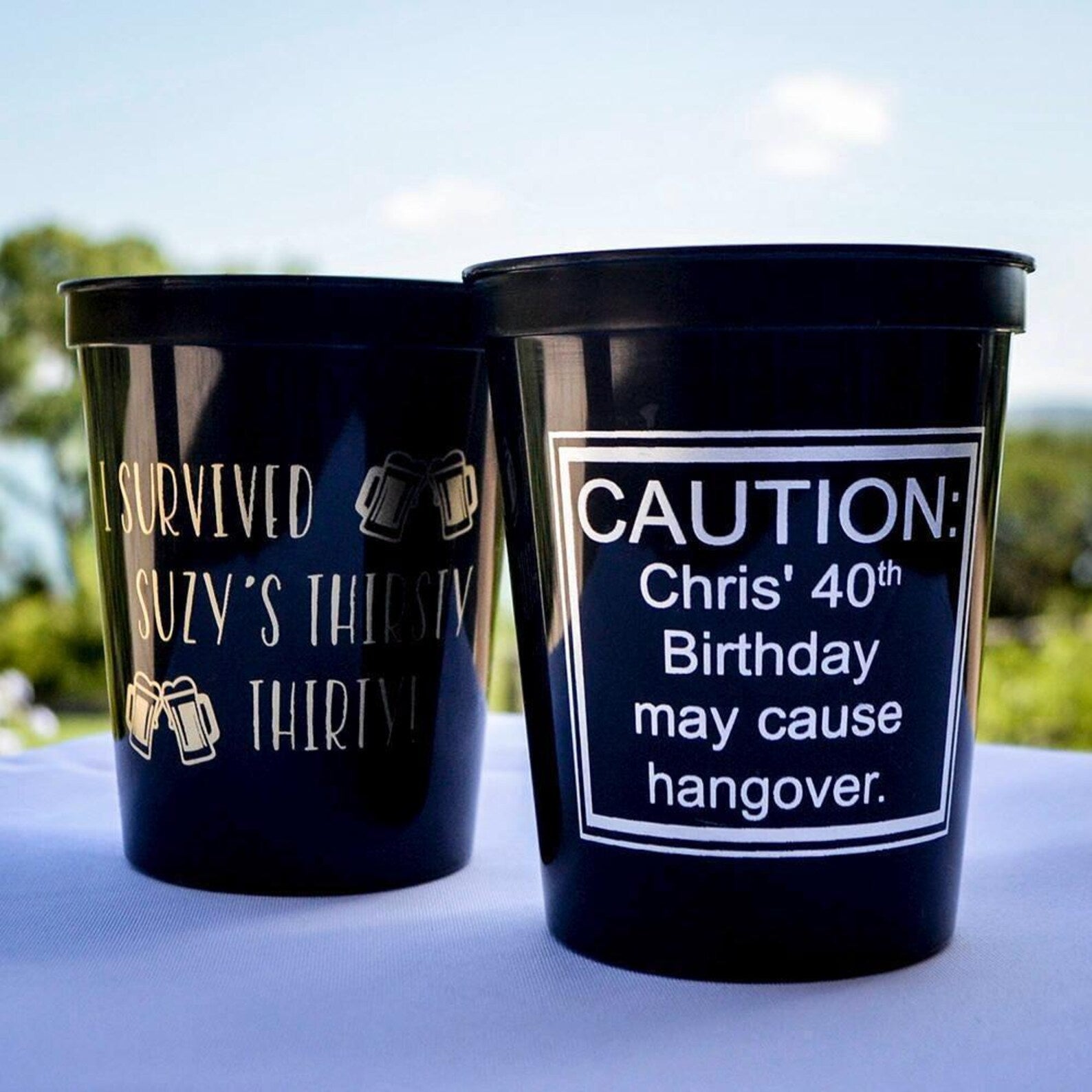 Two black plastic cups with humorous text on them, placed on a table against a blurred outdoor background.