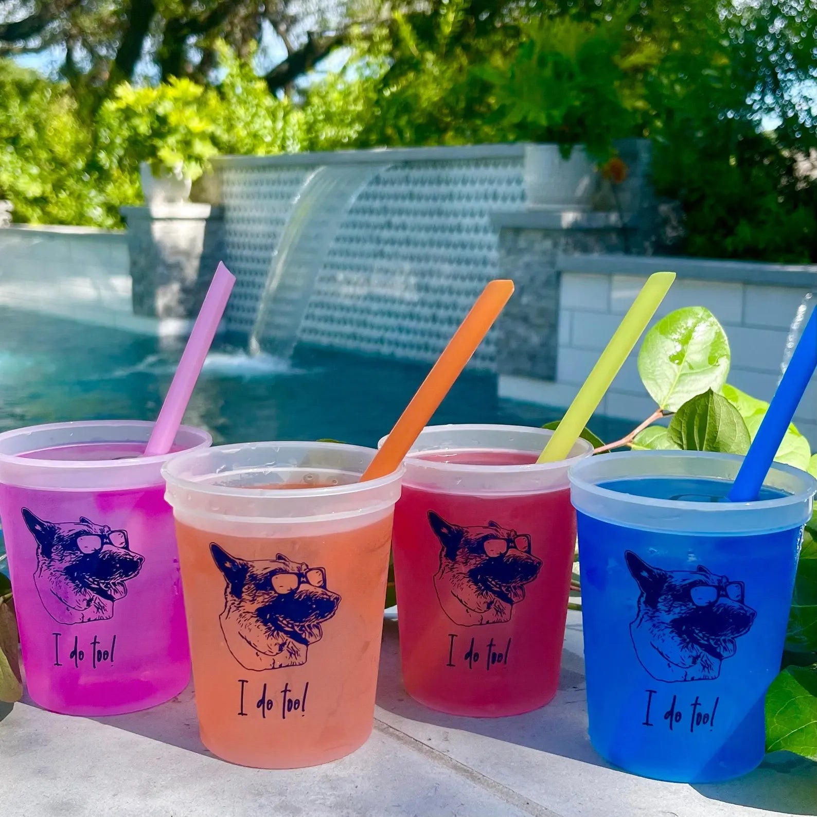 four colorful plastic cups with straws, each containing a different colored liquid, placed on a table near a swimming pool.