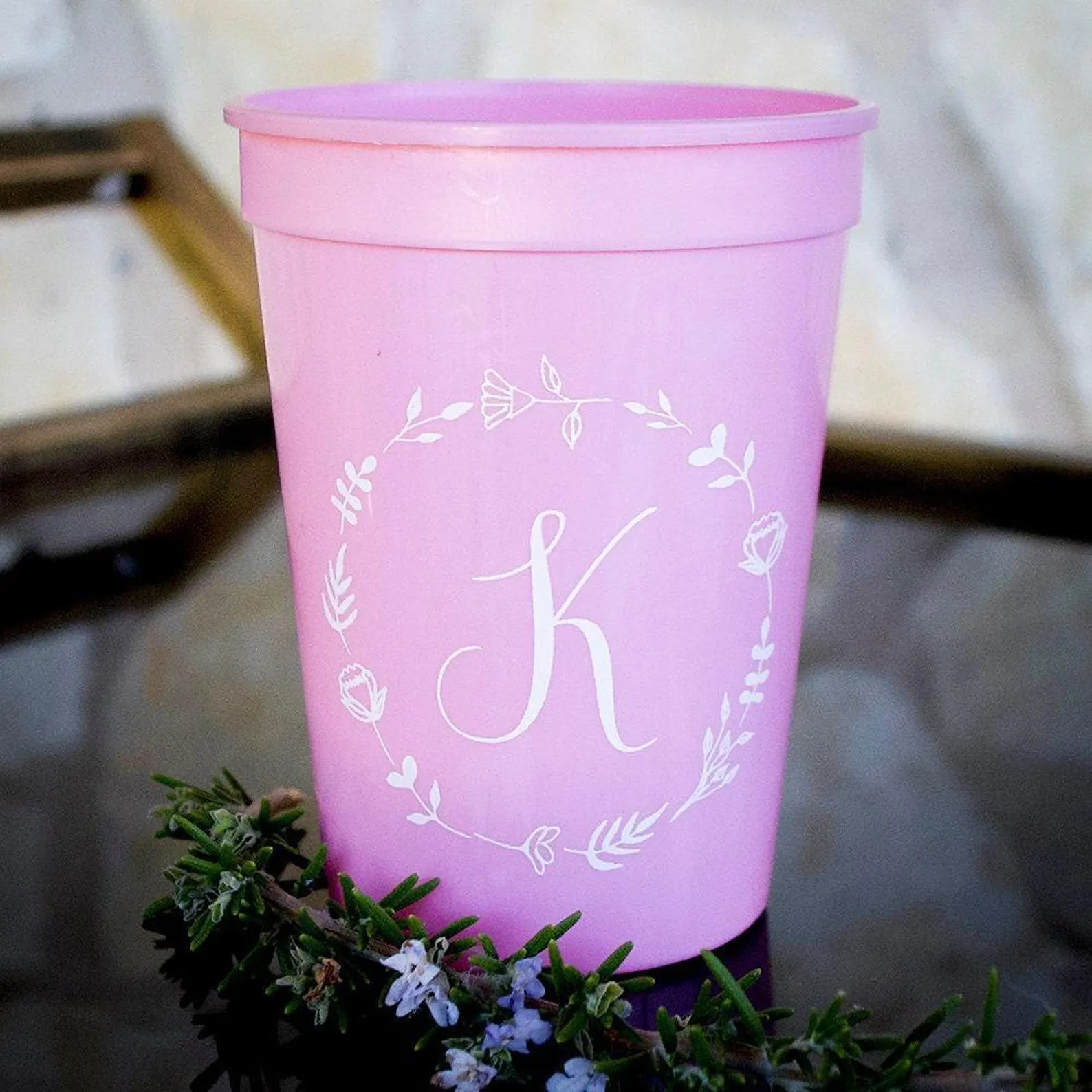 A pink plastic cup with a white monogrammed letter "K" on it, placed on a small green plant or branch.
