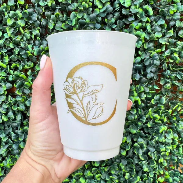 A hand holding a white cup with a gold letter "C" on it, against a background of green leaves.